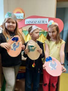 Troje uczniów pozuje do wspólnego zdjęcia trzymając w dłoniach wykonane przez siebie z papieru jaja z dinozaurami. Za dziećmi stoi makieta z napisem "DZIEŃ DINOZAURA".
