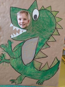 Uczeń pozuje do zdjęcia z makietą przedstawiającą odręcznie namalowanego zielonego dinozaura. Makieta obok oka dinozaura ma okrągły otwór przez, który wystawia głowę uśmiechnięty uczeń.