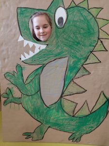 Uczennica pozuje do zdjęcia z makietą przedstawiającą odręcznie namalowanego zielonego dinozaura. Makieta obok oka dinozaura ma okrągły otwór przez, który wystawia głowę uśmiechnięta uczennica.