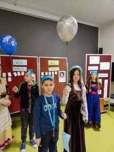 Dwóch uczniów klasy piątej wraz z uczennicami klasy piątej stoją na sali teatralnej. Uczniowie wraz z dziewczętami ustawieni są w dwie pary i trzymają baloniki unoszące się ku górze z przyczepionymi na dole kartkami papieru. W tle widać tablice z punktacją przyznawaną podczas konkursu wiedzy o Mikołaju Koperniku.