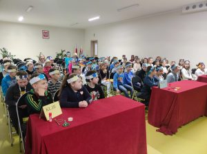 Uczniowie klas I-III siedzą na krzesełkach na sali teatralnej. Dzieci na głowie mają papierowe opaski ozdobione żółtymi i niebieskimi gwiazdkami. Przed uczniami siedzącymi w pierwszym rzędzie ustawiono ławki z numerem klasy, przykryte czerwonym materiałem.