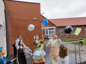 Uczennice klasy piątej wypuszczaja przed budynkiem szkoły kolorowe baloniki. Dziewczynki ubrane sa w stroje z epoki Mikołaja Kopernika. Baloniki przyczepione mają na wstążkach karteczki.