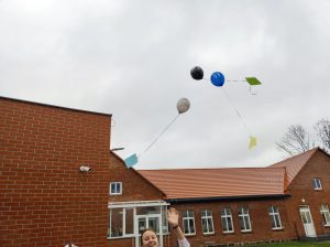 Zdjęcie unoszących się nad budynkiem szkoły trzech baloników z przyczepionymi na wstążkach kolorowymi kartkami.