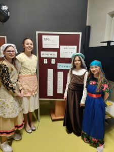 Cztery uczennice klasy piątej pozują przy tablicy z przyczepionymi białymi zadrukowanymi kartkami. Dziewczynki ubrane są w długie suknie z epoki Mikołaja Kopernika. Na tablicy przyczepione są informacje dotyczące wyników w turnieju wiedzy klas I - III takie jak numery zadań i otrzymane w nich punkty przez biorące w nim udział drużyny.