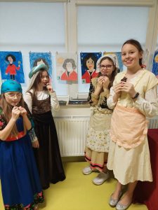 Cztery uczennice klasy piątej jedzą czekoladowe pierniczki. Dziewczynki ubrane sa w długie suknie z epoki Mikołaja Kopernika. W tle za nimi na oknie wisi girlanda utworzona z portretów Mikołaja Kopernika.