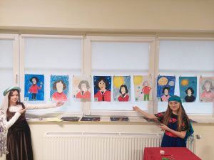 Dwie uczennice klasy piątej stoją przy girlandzie utworzonej z portretów Mikołaja Kopernika. Girlanda zawieszona jest na oknach sali teatralnej. Dziewczynki w strojach z epoki Mikołaja Kopernika wskazują dłońmi na portrety.