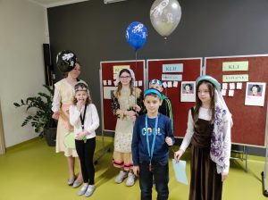 Troje uczniów klas I-III i trzy uczennice klasy piątej stoją parami na sali teatralnej. Każda para trzyma unoszący się w górę balonik. Uczennice klasy piątej ubrane są w stroje z epoki Mikołaja Kopernika a dzieci z klas I-III mają na głowach papierowe opaski. W tle widać tablice z wynikami turnieju wiedzy o Mikołaju Koperniku.