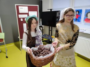 Dwie uczennice klasy piątej trzymają wypełniony czekoladowymi piernikami kosz. Dziewczynki ubrane są w stroje z epoki Mikołaja Kopernika.