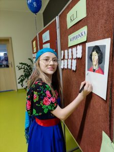 Uczennica klasy piątej stoi i tablicy. Dziewczynka ubrana jest w strój z epoki Mikołaja Kopernika. W dłoni trzyma pisak, który przyciska do przyczepionego do tablicy portretu Mikołaja Kopernika.