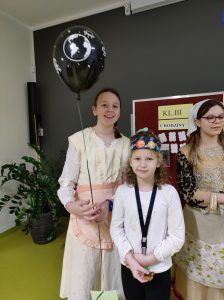 Do zdjęcia pozują dwie uczennice - jedna z klas I-III, druga z klasy piątej. Starsza dziewczynka trzyma wstążeczkę do której przyczepiony jest czarny balonik.Młodsza dziewczynka na głowie ma papierowa opaskę z grafiką planet układu słonecznego. Obok dziewczynek stoi uczennica klasy piątej ubrana w strój z epoki Mikołaja Kopernika.