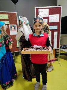 Uczennica klasy trzeciej stoi z tacą wypełnioną czekoladowymi pierniczkami. Dziewczynka na głowie ma papierową opaskę z grafikami planet układu słonecznego. W tle widać odwrócone tyłem uczennice klasy piątej ubrane w stroje z epoki Mikołaja Kopernika.