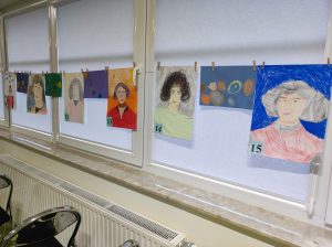 Zdjęcie zawieszonej na oknie sali gimnastycznej girlandy. Na girlandzie przy pomocy klamerek zawieszono portrety Mikołaja Kopernika wykonane przez uczniów. Portrety w lewym dolnym rogu przyklejone mają karteczki z numerkami.