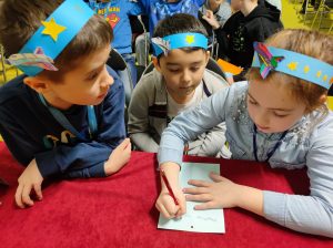 Troje uczniów klasy trzeciej pochyla się nad karteczką leżąca na przykrytym czerwonym materiałem stoliku. Dziewczynka siedząca po prawej stronie trzyma w dłoni długopis i pisze cos na karteczce. Dzieci na głowach maja niebieskie papierowe opaski z przyklejonymi rysunkami statków kosmicznych i gwiazdek.
