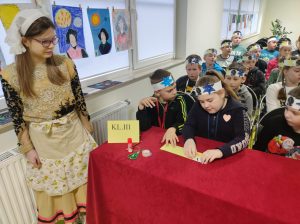 Uczniowie klasy trzeciej siedzą w rzędach. Troje uczniów z pierwszego rzędu siedzi za stolikiem przykrytym czerwonym materiałem. Na stoliku stoi karteczka napisem kl. III oraz leży podłużna koperta, której przygląda się siedząca po środku uczennica. Dzieci na głowach mają papierowe opaski z przyczepionymi gwiazdkami. Obok nich stoi uczennica klasy piątej w stroju z epoki Mikołaja kopernika.