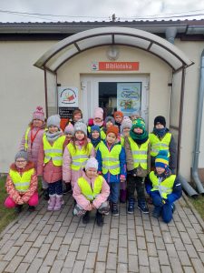 Uczniowie klasy pierwszej pozują do wspólnego zdjęcia przed drzwiami do biblioteki w Ostaszewie. Nad drzwiami wisi czerwona tabliczka z napisem "Biblioteka". Dzieci ubrane są w odblaskowe kamizelki.