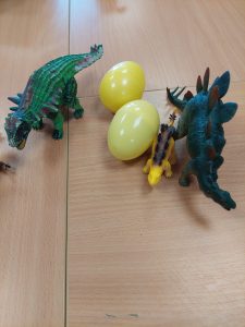 Zdjęcie trzech figurek dinozaurów ustawionych obok siebie. Pomiędzy dinozaurami leżą dwa żółte, plastikowe jajka.