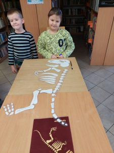 Dwoje uczniów klasy pierwszej stoi na szczycie podłużnego stołu na którym leży biały szkielet dinozaura, wycięty z papieru oraz czerwona kartka papieru z mniejszym szkieletem..