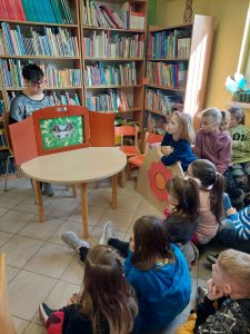 Uczniowie klasy pierwszej podczas oglądania teatrzyku w gminnej bibliotece. Na przeciwko uczniów ustawiona jest mała scena, za którą siedzi Pani bibliotekarka.