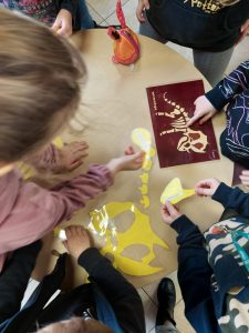 Uczniowie klasy pierwszej pochylają się nad stołem, na którym leżą zalaminowane fragmenty szkieletu dinozaura oraz czerwona kartka z odciśniętym na żółto pełnym szkieletem. Uczniowie oglądają fragmenty szkieletu i biorą je w ręce.