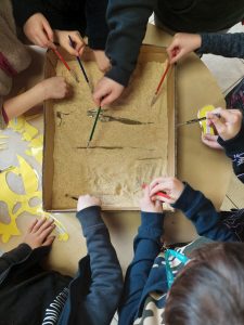 Uczniowie klasy pierwszej pochylają się nad kartonem wypełnionym piaskiem. Dzieci trzymają w dłoniach pędzelki, którymi robią ślady na piasku.