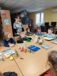 Uczniowie klasy pierwszej siedzą przy dużym stole w bibliotece w Ostaszewie. Na stole leżą porozkładane książki i figurki z dinozaurami. Obok uczniów stoi Pani Bibliotekarka.