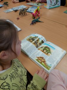 Uczennica klasy pierwszej siedzi przy stoliku. Przed nią leży rozłożona książka ze zdjęciami dinozaura. W tle widać leżące na stole inne książki oraz figurki dinozaurów.
