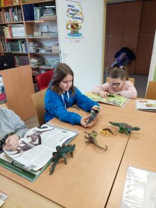 Dwie uczennice oddziału przedszkolnego siedzą przy dużym stole. Na stole leżą figurki dinozaurów oraz rozłożone książki. Jedna z uczennic z uwagą czyta książkę, a druga ogląda figurkę dinozaura.