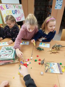 Dzieci siedzą przy dużym stole. Na stole porozrzucane są książki i kolorowe małe pomponiki. Uczniowie wykonują swoje kartki z dinozaurami. Na zdjęciu trzy uczennice podczas wykonywania swojej pracy.
