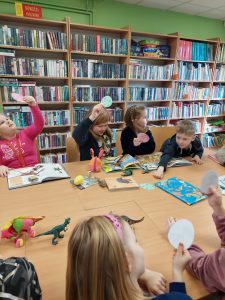 Uczniowie klasy pierwszej siedzą przy dużym stole w bibliotece publicznej. Na stole leżą otwarte książki i figurki z dinozaurami. Dzieci podnoszą w górę papierowa koła, którym się przyglądają.