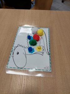 Zdjęcie wsadzonej w folię kartki z nadrukowanym konturem dinozaura. Dinozaur ozdobiony jest małymi, kolorowymi pomponami. Całość kartki ozdobiona jest zieloną ramką utworzoną z przerywanych linii.