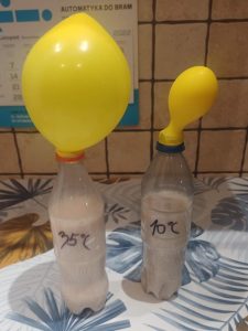 Na stole ustawiono dwie plastikowe butelki częściowo wypełnione białym płynem. Na butelce po lewej stronie odręcznie napisano 35 stopni C a na prawej 10 stopni C. Na obie butelki od góry naciągnięto żółte balony. Balonik na butelce po lewej stronie jest nadmuchany i unosi się do góry, a ten po prawej stronie jest dużo mniejszy.