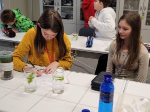 Dwie uczennice siedzą przy stoliku. Przed nimi stoją dwie szklane menzurki wypełnione częściowo wodą, w których pływają kawałki roślin. Jedna z dziewczyn robi notatkę na kartce. Druga się jej przygląda. W tle widać innych pracujących uczniów.