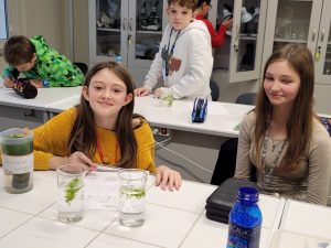 wie uczennice siedzą przy stoliku. Przed nimi stoją dwie szklane menzurki wypełnione częściowo wodą, w których pływają kawałki roślin. Dziewczynki się uśmiechają. W tle widać innych pracujących uczniów.