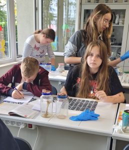 Przy stoliku siedzi dwoje uczniów klasy siódmej. Przed nimi stoją słoiki , w których widać pojedyncze kości. W tle stoją dwie uczennice. Jedna pochyla się nad stolikiem.