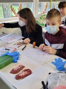 Dwoje uczniów klasy siódmej z maseczkami na twarzy siedzi przy stoliku. Przed nimi leżą dwie nerki. Wokół nich na stole porozkładane są przybory do pisania oraz niebieskie, jednorazowe rękawiczki.