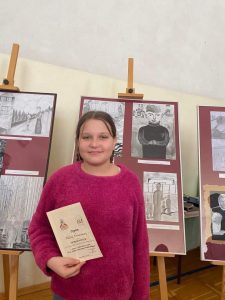 Uczennica Paulina Chrzanowska pozuje do zdjęcia z otrzymanym dyplomem. Dziewczynka lekko się uśmiecha. Stoi na tle nagrodzonych prac.