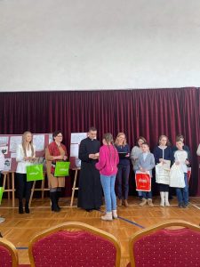 Uczennica Paulina Chrzanowska odbiera z rąk księdza nagrodę. Obok księdza stoją nagrodzeniu już uczniowie oraz Pani trzymające zielone torby z upominkami.