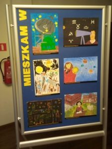 Zdjęcie niebieskiej tablicy na której pionowo umieszczono żółty napis "MIESZKAM W" a obok niego 6 prac plastycznych wyróżnionych w konkursie.