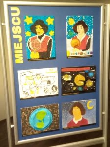 Zdjęcie niebieskiej tablicy na której pionowo umieszczono żółty napis "MIEJSCU" a obok niego 6 prac plastycznych wyróżnionych w konkursie.