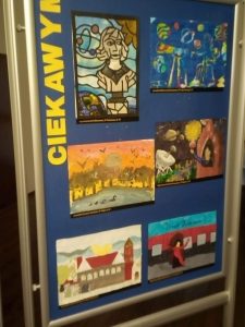 Zdjęcie niebieskiej tablicy na której pionowo umieszczono żółty napis "CIEKAWYM" a obok niego 6 prac plastycznych wyróżnionych w konkursie.