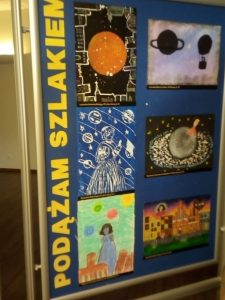 Zdjęcie niebieskiej tablicy na której pionowo umieszczono żółty napis "PODĄŻAM SZLAKIEM" a obok niego 6 prac plastycznych wyróżnionych w konkursie.
