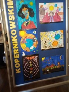 Zdjęcie niebieskiej tablicy na której pionowo umieszczono żółty napis "KOPERNIKOWSKIM" a obok niego 6 prac plastycznych wyróżnionych w konkursie.