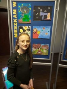 Nagrodzona w konkursie uczennica Laura Kontowicz pozuje do zdjęcia na tle galerii stworzonej z nagrodzonych prac plastycznych.