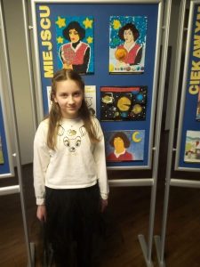 Nagrodzona w konkursie uczennica Wiktoria Abramowicz pozuje do zdjęcia na tle galerii stworzonej z nagrodzonych prac plastycznych.