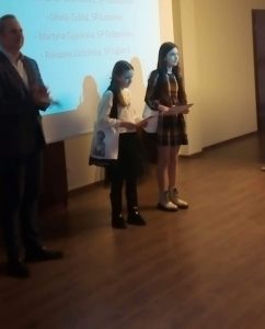 Dwie nagrodzone w konkursie uczennice naszej szkoły pozują do wspólnego zdjęcia. dziewczynki trzymają w dłoniach dyplomy i stoją na tle ekranu projekcyjnego, na którym wyświetlone są nazwiska zwycięzców w kategorii klas IV-VI.