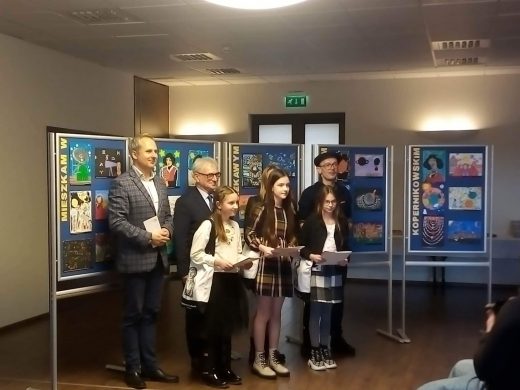 Dwie uczennice naszej szkoły nagrodzone w konkursie pozują do wspólnego zdjęcia z organizatorami konkursu. Wszyscy stoją na tle galerii nagrodzonych w konkursie prac.