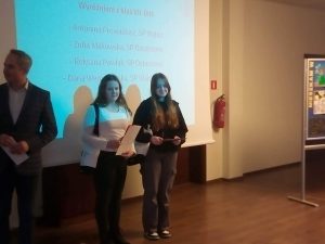 Zdjęcie dwóch wyróżnionych w konkursie uczennic naszej szkoły. Dziewczyny stoją na tle ekranu projekcyjnego, na którym wyświetlają się imiona i nazwiska wyróżnionych w konkursie uczniów klas VII-VIII.