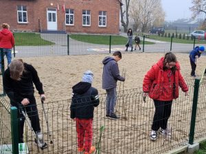 Uczniowie klasy czwartej chodzą po piaskownicy szkolnego placu zabaw. Dzieci oglądają wbite w piasek tyczki tworzące stację meteoroliczną.