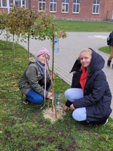 Dwie uczennice klasy czwartej kucają na trawniku przed szkołą. przy wbitych w ziemię tyczkach tworzących stację meteorologiczną.