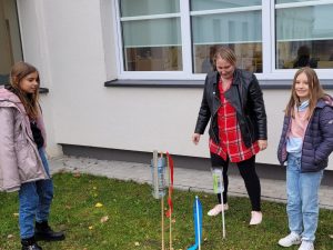 Dwie uczennice wraz z nauczycielką stoją na trawniku przed budynkiem szkoły. Przed nimi w ziemi umieszczone są tyczki z umieszczonymi u góry połówkami plastikowych butelek, tworzące stację meterologiczną.